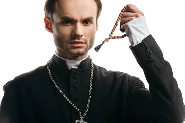 Young, serious catholic priest holding wooden rosary beads while looking at camera isolated on white — Stock Photo