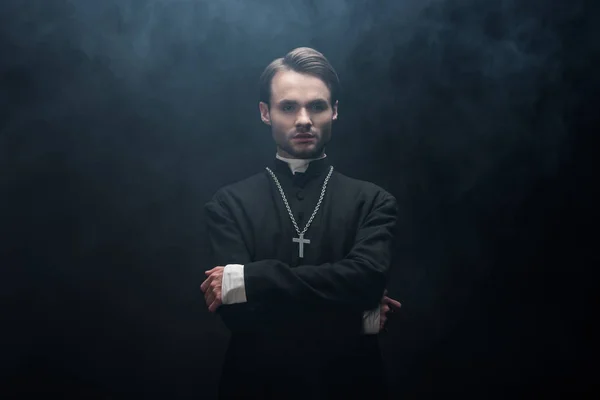 Young serious catholic priest standing with crossed arms on black background with smoke — Stock Photo