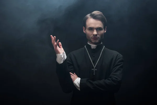 Young confident catholic priest standing with open arm on black background with smoke — Stock Photo