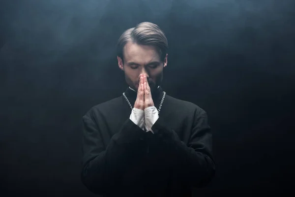 Joven sacerdote católico concentrado rezando con los ojos cerrados sobre fondo negro con humo - foto de stock