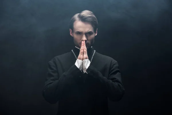 Serio sacerdote cattolico guardando la macchina fotografica mentre prega su sfondo nero con fumo — Foto stock