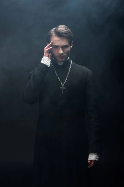 Young thoughtful catholic priest touching head while looking at camera on black background with smoke — Stock Photo