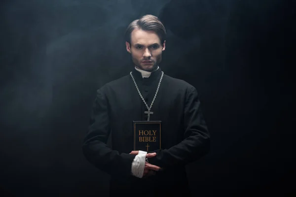 Young confident catholic priest looking at camera while holding holy bible on black background with smoke — Stock Photo