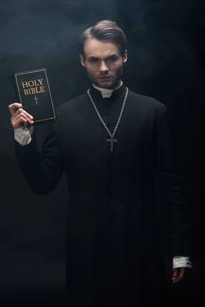 Young strict catholic priest showing holy bible at camera on black background with smoke — Stock Photo