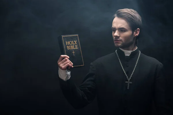 Thoughtful catholic priest looking at holy bible on black background with smoke — Stock Photo