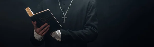 Partial view of catholic priest reading holy bible isolated on black, panoramic shot — Stock Photo