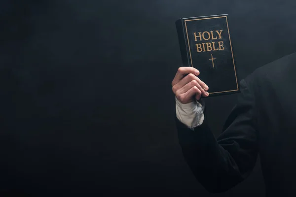 Cropped view of catholic priest holding holy bible isolated on black — Stock Photo