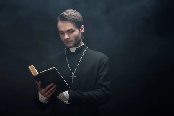 Attentive catholic priest reading holy bible on black background with smoke — Stock Photo