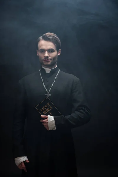 Confident catholic priest holding holy bible and looking at camera on black background with smoke — Stock Photo