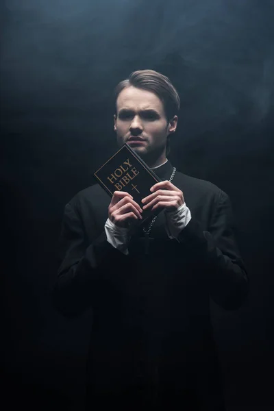 Tense catholic priest looking at camera while holding holy bible on black background with smoke — Stock Photo