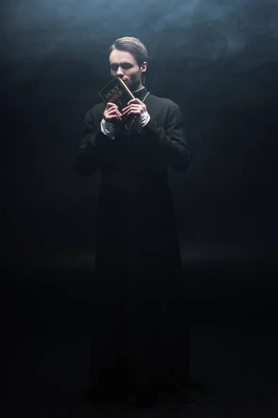 Full length view of young catholic priest kissing holy bible on black background with smoke — Stock Photo