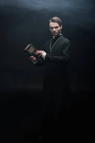 Full length view of strict catholic priest holding holy bible and looking at camera on black background with smoke — Stock Photo