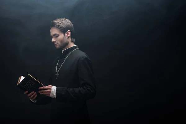 Prêtre catholique concentré lisant la bible sainte sur fond noir avec de la fumée — Photo de stock