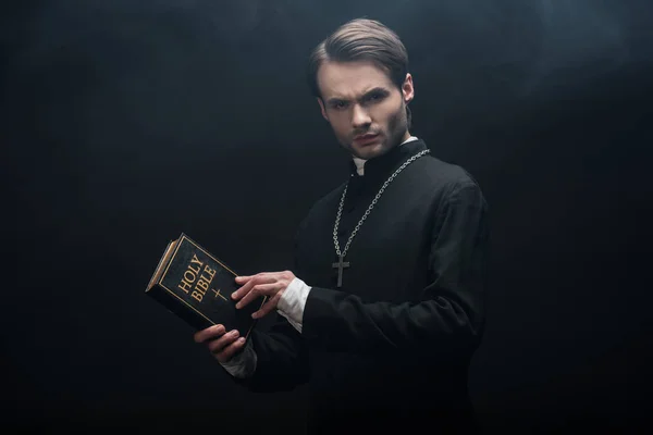Strict catholic priest holding holy bible and looking at camera on black background with smoke — Stock Photo
