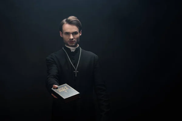 Young serious catholic priest holding holy bible in outstretched hand isolated on black — Stock Photo