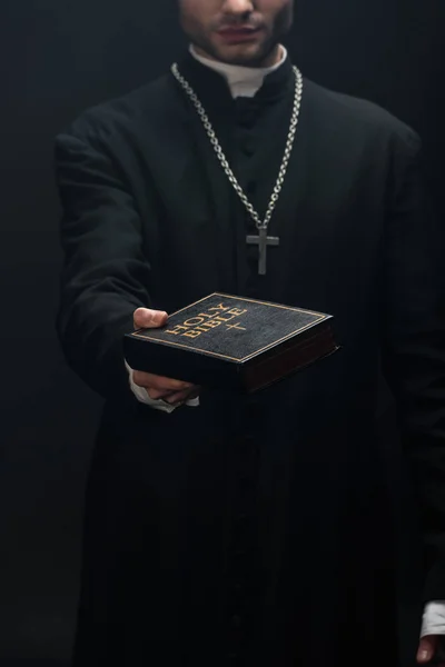 Cropped view of catholic priest holding holy bible in outstretched hand isolated on black — Stock Photo