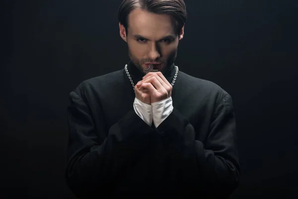 Joven sacerdote católico reflexivo sosteniendo cruz de plata en su collar aislado en negro - foto de stock