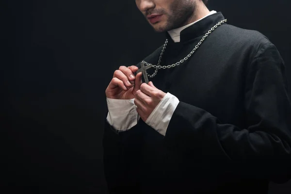 Vista cortada de padre católico olhando para cruz de prata em seu colar isolado em preto — Fotografia de Stock