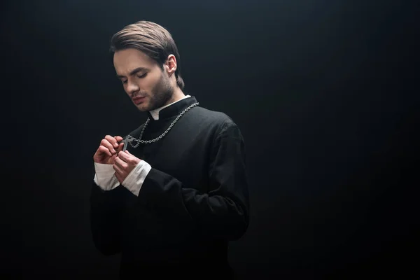 Young serious catholic priest looking at silver cross on his necklace isolated on black — Stock Photo