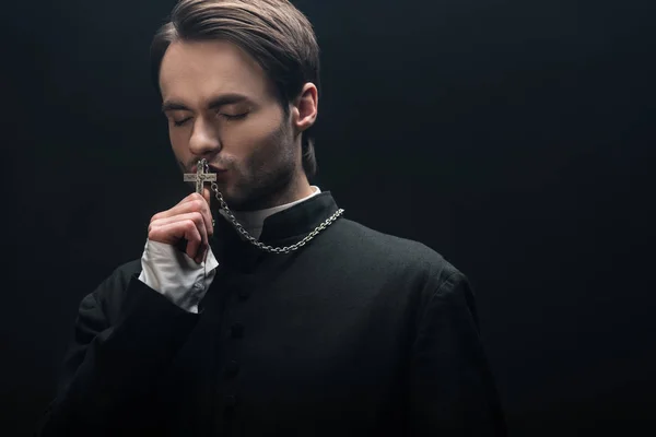 Young thoughtful catholic priest kissing silver cross with closed eyes isolated on black — Stock Photo