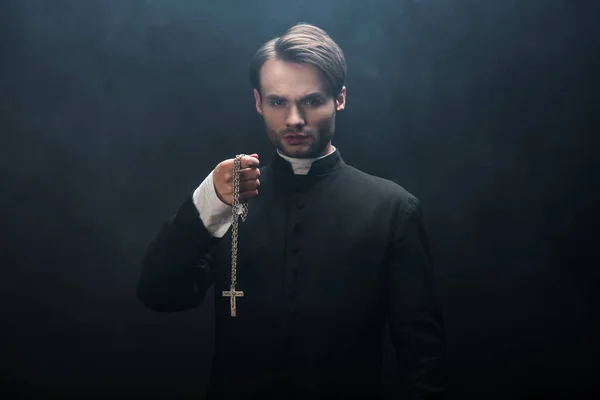 Confident catholic priest holding necklace with cross while looking at camera on black background with smoke — Stock Photo