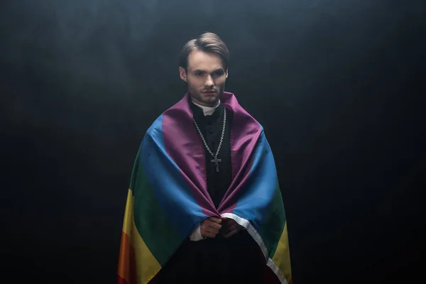 Serious catholic priest wrapped in lgbt flag while looking at camera on black background with smoke — Stock Photo