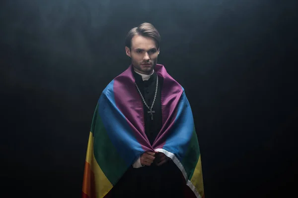 Sacerdote católico grave envolto em lgbt bandeira enquanto olha para a câmera no fundo preto com fumaça — Fotografia de Stock