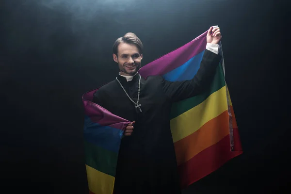 Sorrindo padre católico segurando bandeira lgbt enquanto olha para a câmera no fundo preto com fumaça — Fotografia de Stock