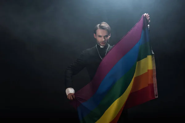 Впевнений католицький священик тримає прапор lgbt, дивлячись на камеру на чорному тлі з димом — стокове фото