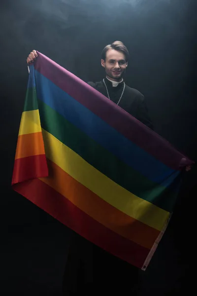Sorrindo padre católico segurando bandeira lgbt enquanto olha para a câmera no fundo preto com fumaça — Fotografia de Stock