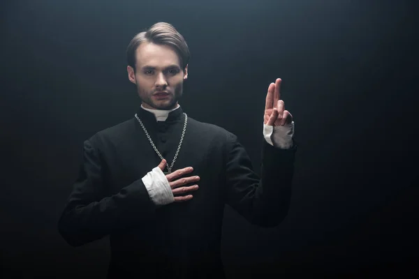 Confident catholic priest showing blessing gesture and holding hand on chest isolated on black — Stock Photo
