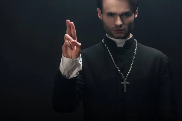 Young confident catholic priest looking at camera and showing blessing gesture isolated on black — Stock Photo