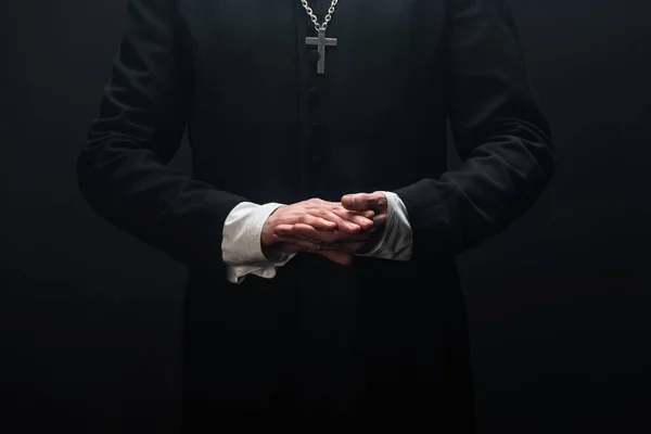 Cropped of catholic priest standing with folded hands isolated on black — Stock Photo