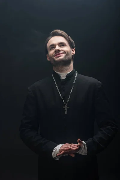 Padre católico feliz de pé com as mãos dobradas e olhando para cima isolado em preto — Fotografia de Stock
