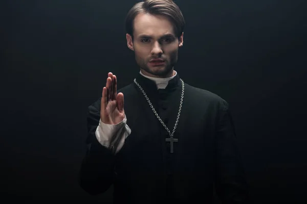 Молодий впевнений католицький священик дивиться на камеру і демонструє благословенний жест ізольовано на чорному — стокове фото