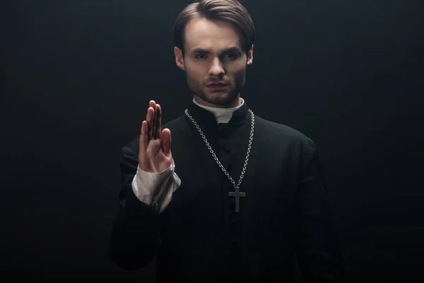 Young confident catholic priest looking at camera and showing blessing gesture isolated on black — Stock Photo