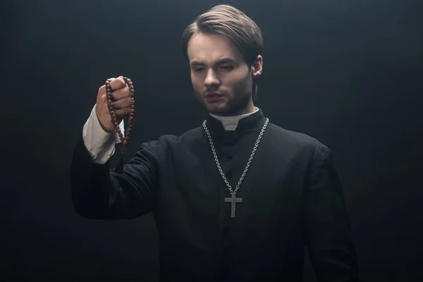 Young tense catholic priest looking at wooden rosary beads isolated on black — Stock Photo