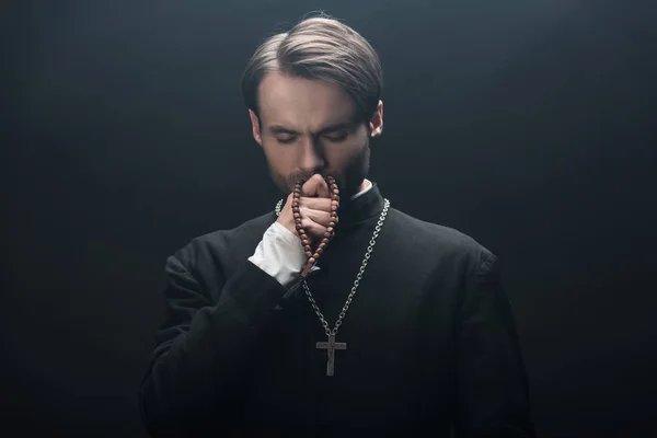 Pensativo sacerdote católico besar cruz de plata con los ojos cerrados aislados en negro - foto de stock