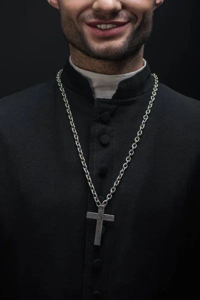 Abgeschnittene Ansicht eines lächelnden katholischen Priesters mit silbernem Kreuz an der Kette isoliert auf schwarz — Stockfoto