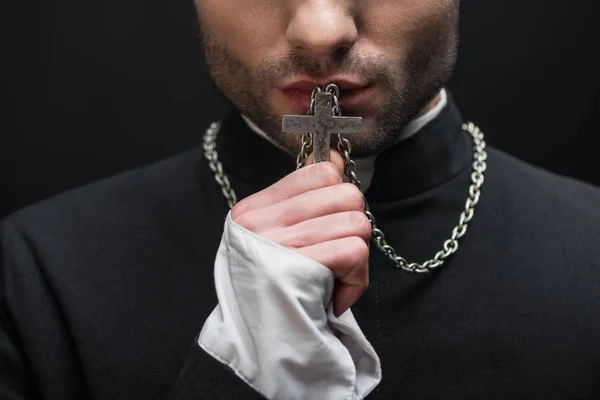 Обрезанный вид католического священника целующего серебряный крест на своем ожерелье изолирован от черного — стоковое фото