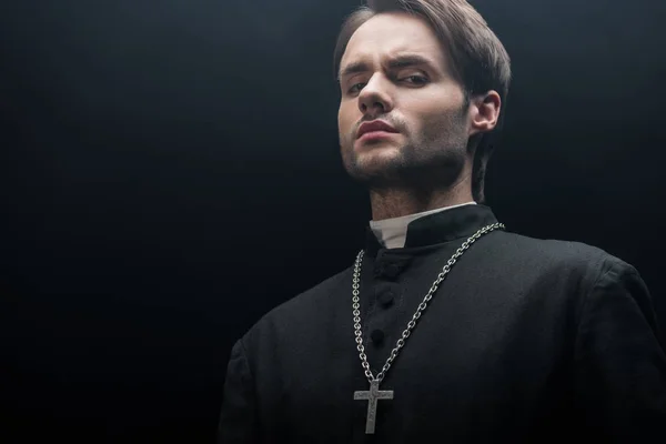 Vista de ángulo bajo del joven sacerdote católico tenso mirando a la cámara aislada en negro - foto de stock