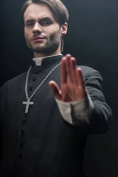 Vista de ángulo bajo del sacerdote católico estricto mostrando gesto de basura aislado en negro - foto de stock