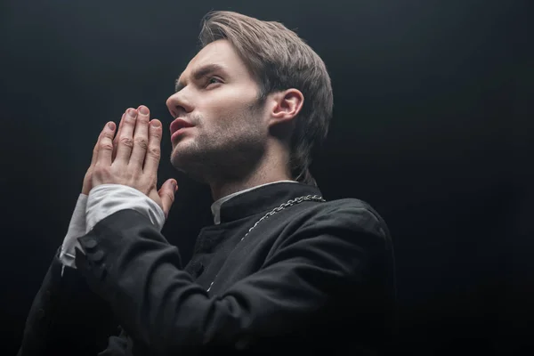 Low angle veiw of young concentrated catholic priest praying isolated on black — Stock Photo