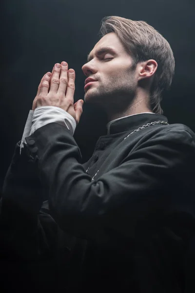 Veta de ángulo bajo de joven sacerdote católico concentrado orando con los ojos cerrados aislados en negro - foto de stock