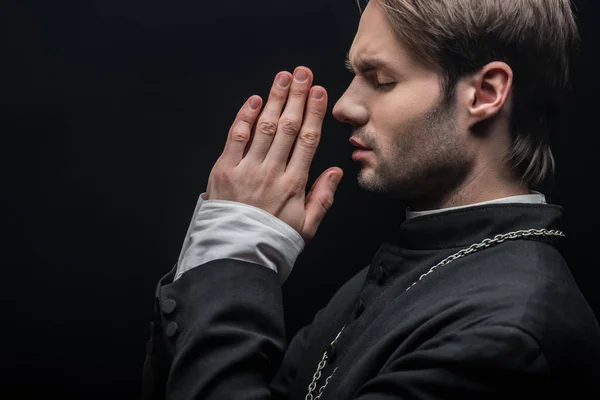 Vista lateral del joven sacerdote católico concentrado orando con los ojos cerrados aislados en negro - foto de stock