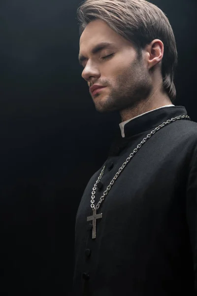 Young catholic priest standing in darkness with closed eyes isolated on black — Stock Photo