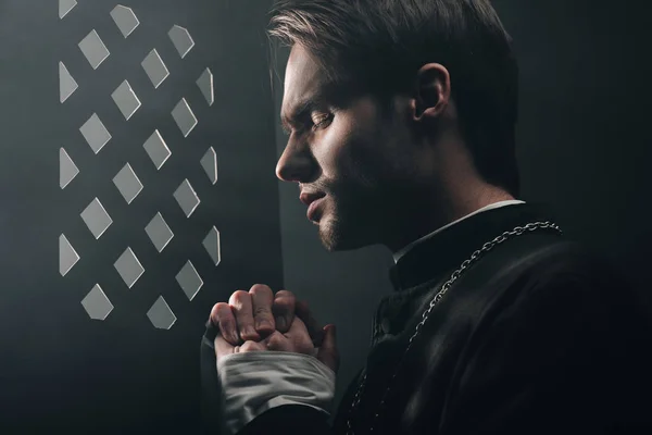 Young thoughtful catholic priest praying with closed eyes in dark near confessional grille with rays of light — Stock Photo