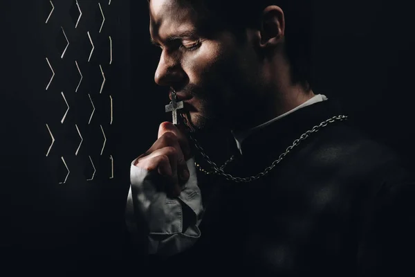 Young tense catholic priest kissing cross on his necklace in dark near confessional grille with rays of light — Stock Photo