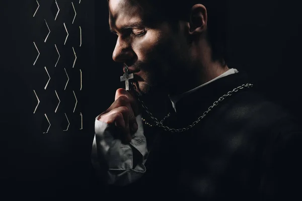 Jovem padre católico tenso beijando cruz em seu colar no escuro perto da grade confessional com raios de luz — Fotografia de Stock