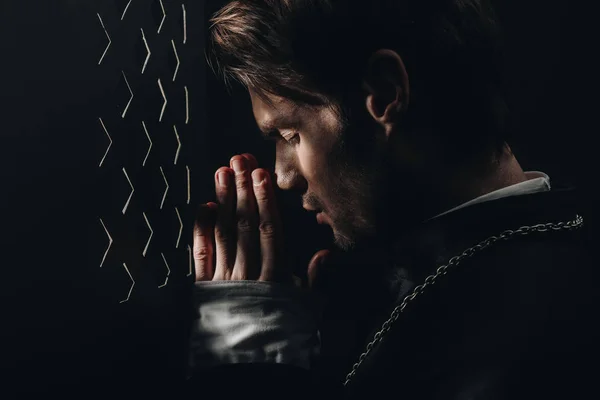 Young catholic priest praying with closed eyes near confessional grille in dark with rays of light — Stock Photo